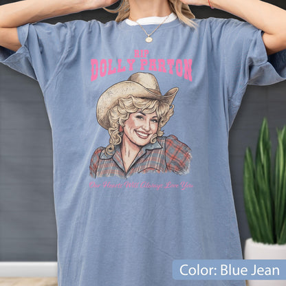 May include: A blue jean colored t-shirt featuring a graphic illustration of Dolly Parton wearing a cowboy hat and a plaid shirt. The text above reads RIP DOLLY PARTON in pink, and the text below reads Our Hearts Will Always Love You.