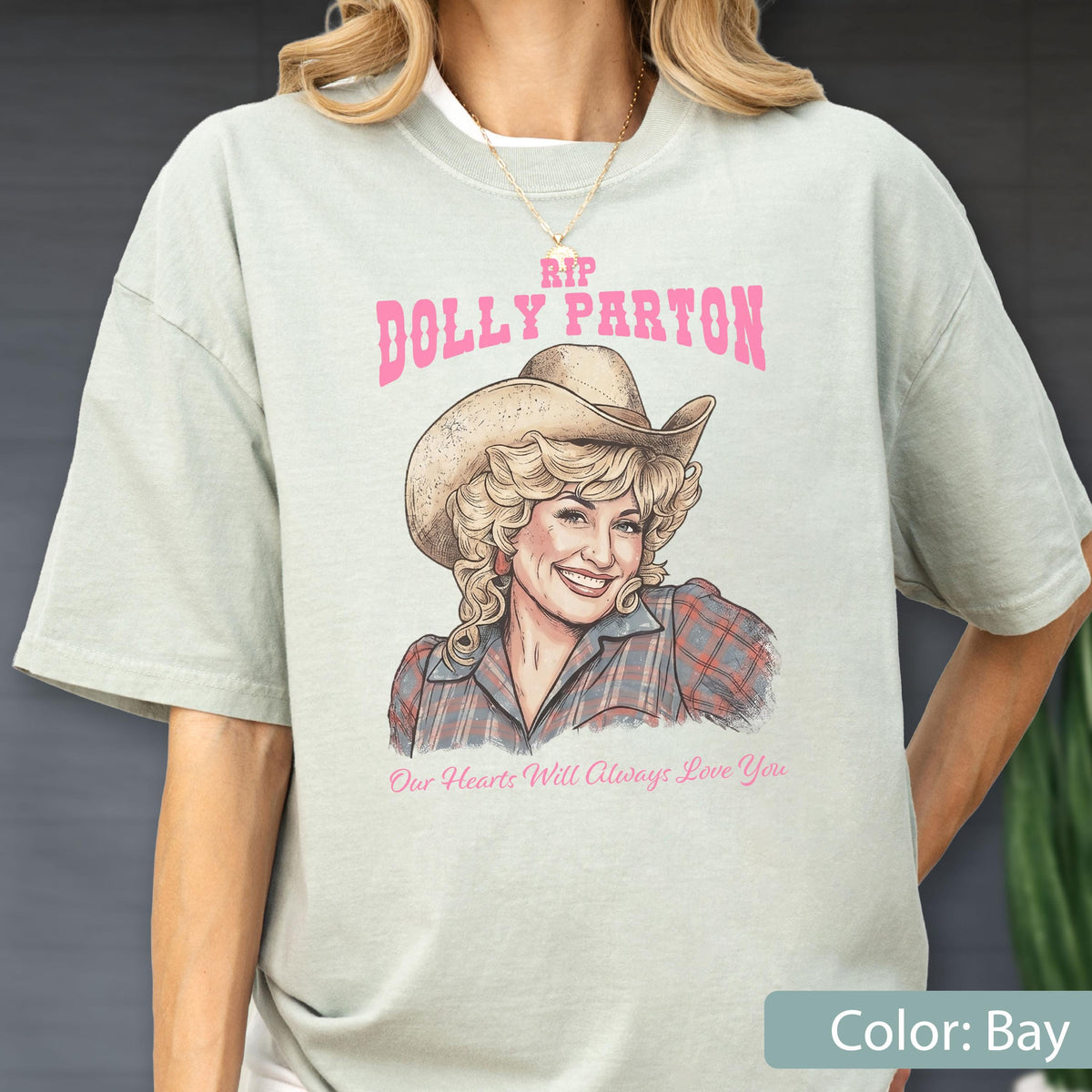 May include: A light sage green t-shirt featuring a graphic illustration of Dolly Parton wearing a cowboy hat and a plaid shirt. The text above reads RIP DOLLY PARTON in pink, and the text below reads Our Hearts Will Always Love You in pink.