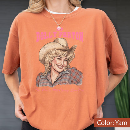 May include: A burnt orange short-sleeve t-shirt featuring a graphic print of Dolly Parton wearing a cowboy hat and a plaid shirt. The text DOLLY PARTON appears in pink above the illustration, with additional text printed below the image.