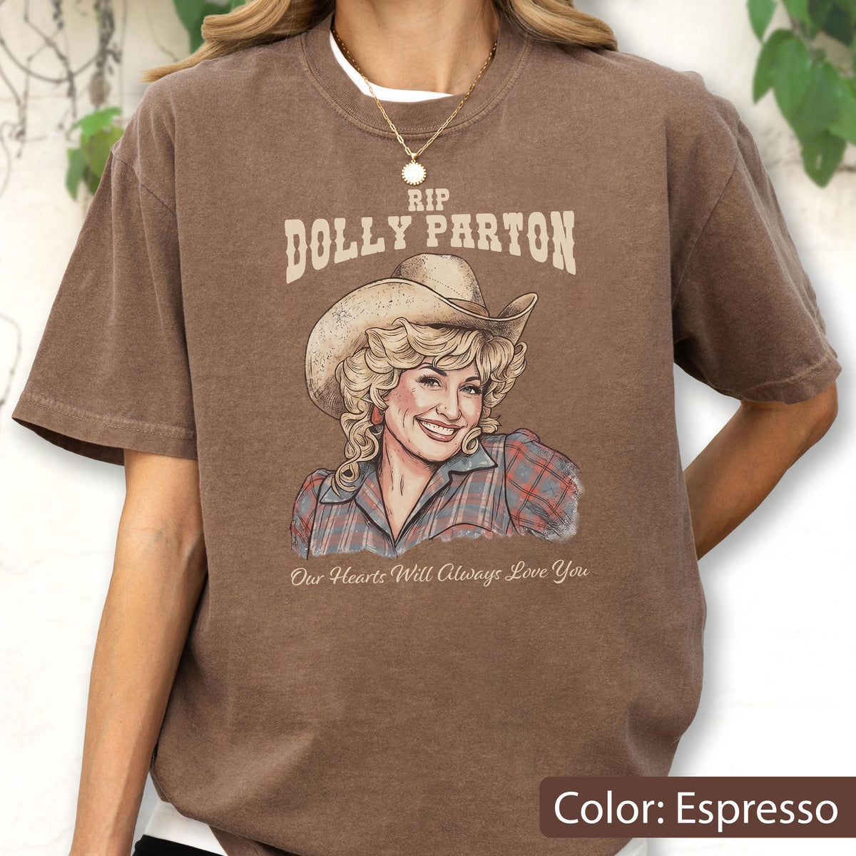 May include: An espresso brown t-shirt featuring a graphic illustration of Dolly Parton wearing a cowboy hat and a plaid shirt. The text above reads RIP DOLLY PARTON and the text below reads Our Hearts Will Always Love You in a script font.