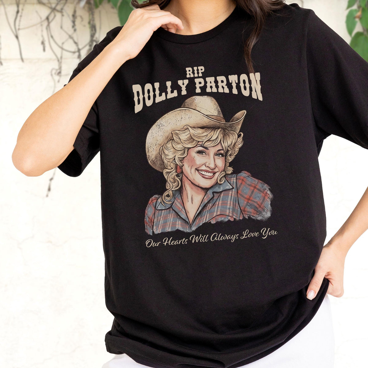 May include: A black short-sleeved t-shirt featuring a graphic print of Dolly Parton wearing a cowboy hat and a plaid shirt. The text above reads RIP DOLLY PARTON and the text below reads Our Hearts Will Always Love You in white lettering.