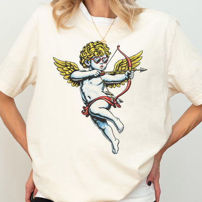 May include: Cream-colored t-shirt featuring a graphic of Cupid with yellow wings, curly hair, and heart-shaped sunglasses, holding a bow and arrow. The design is in a vintage illustration style.