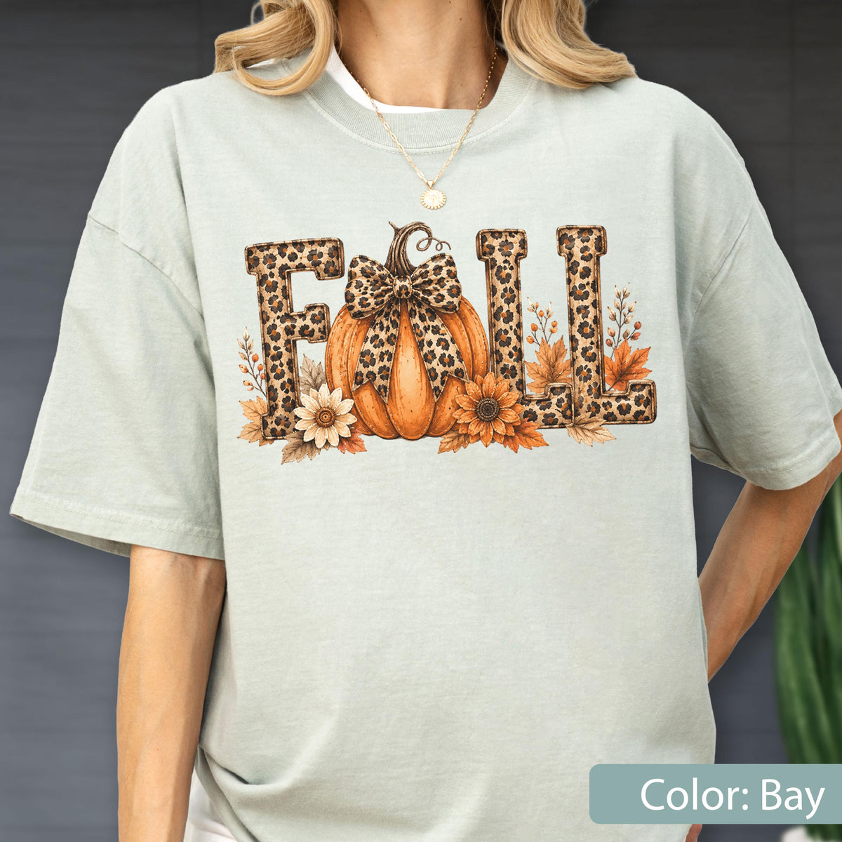 May include: A light sage green t-shirt featuring a graphic print of the word FALL in a leopard print pattern. The letter A is replaced by an orange pumpkin with a leopard print bow, surrounded by orange and yellow autumn leaves and flowers.