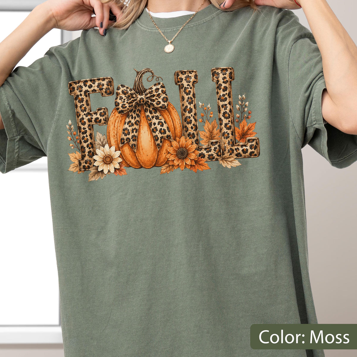 May include: A moss green t-shirt featuring the word FALL in leopard print lettering. The letter A is replaced by an orange pumpkin adorned with a leopard print bow, surrounded by sunflowers and autumn leaves in shades of brown and gold.
