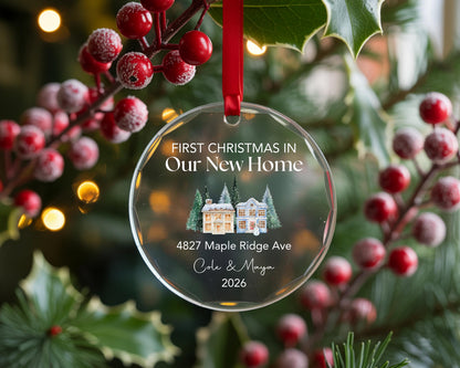 May include: A clear, circular glass ornament hanging from a red ribbon. It features the text "FIRST CHRISTMAS IN Our New Home", "4827 Maple Ridge Ave", "Cole & Maya", and "2026" above and below a watercolor illustration of two houses and pine trees.