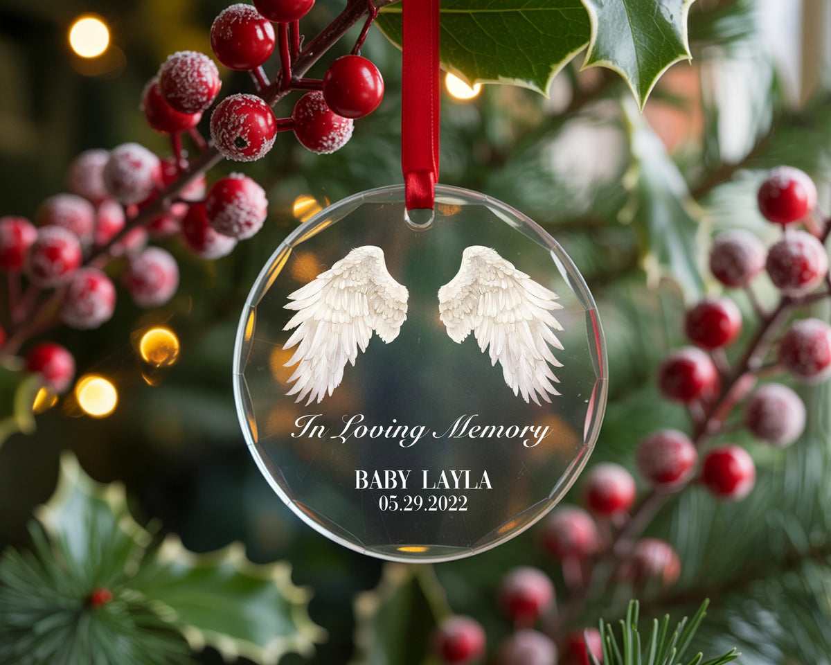 May include: A clear, circular glass ornament hanging from a red ribbon. The ornament features white angel wings printed in the center, with the text In Loving Memory, BABY LAYLA, and the date 05.29.2022 printed in white below the wings.