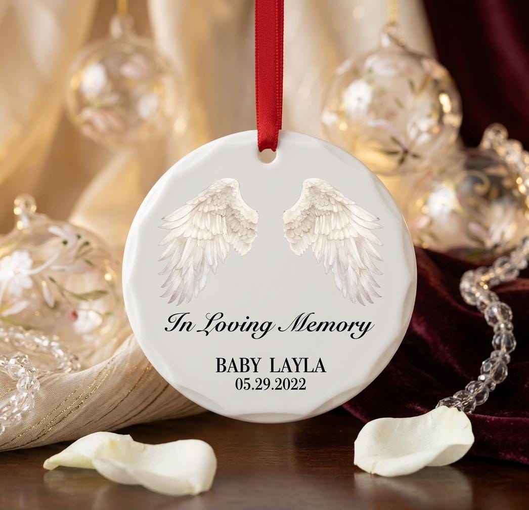 May include: A round white ceramic ornament featuring a pair of detailed white angel wings printed at the top. The text reads In Loving Memory, BABY LAYLA, and 05.29.2022 in black script and serif fonts. A red ribbon is attached for hanging.