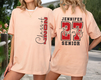 May include: A peach-colored t-shirt featuring a vertical black and red graphic on the front that reads Class of 2027. The back displays the name Jennifer above the number 27 in red with black outlines, the word twenty, and the word Senior.