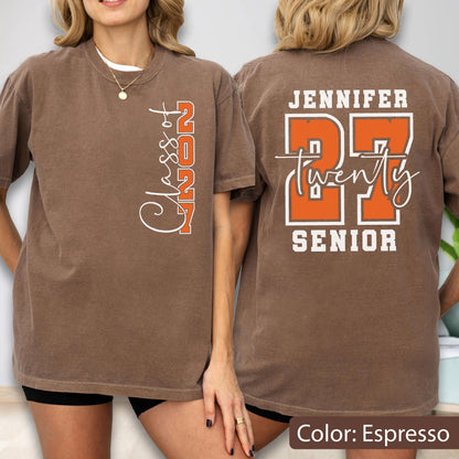 May include: An espresso-colored short-sleeve t-shirt featuring a vertical white and orange graphic on the front that reads Class of 2027. The back displays the name Jennifer in white, the large orange number 27, the word twenty in script, and the word SENIOR.