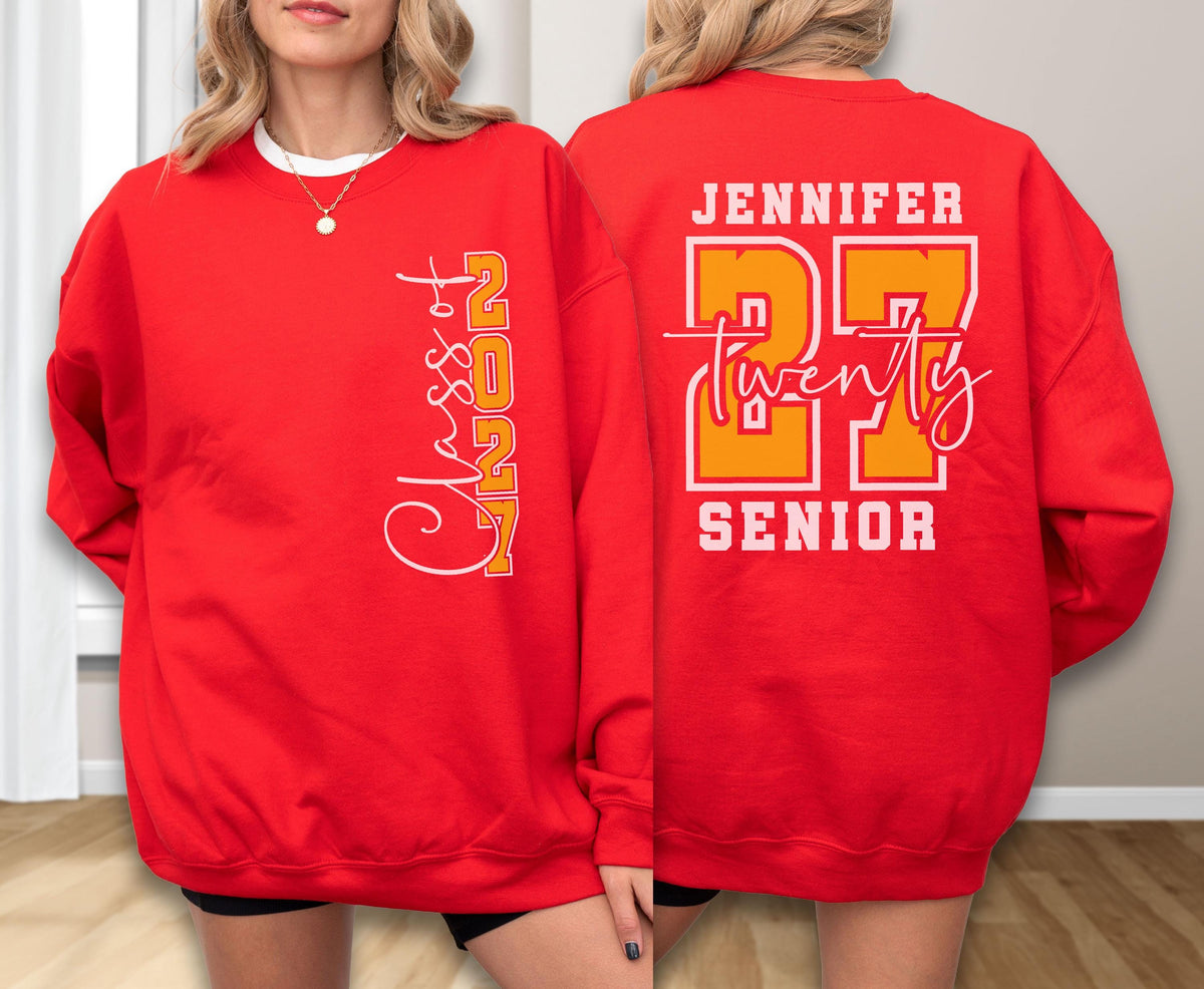 May include: A bright red crewneck sweatshirt featuring white and orange graphic text. The front displays vertical text reading Class of 2027. The back features the name Jennifer, the number 27 in large orange font, the word twenty in script, and Senior.