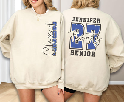 May include: A beige crewneck sweatshirt featuring a blue and black graphic design. The front displays vertical text reading Class of 2027. The back features the name Jennifer, the number 27, the word twenty in script, and the word SENIOR in block letters.