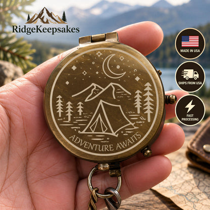A hand holding a compass with a design of mountains, trees, and a tent, with the text "Adventure Awaits" printed on it.