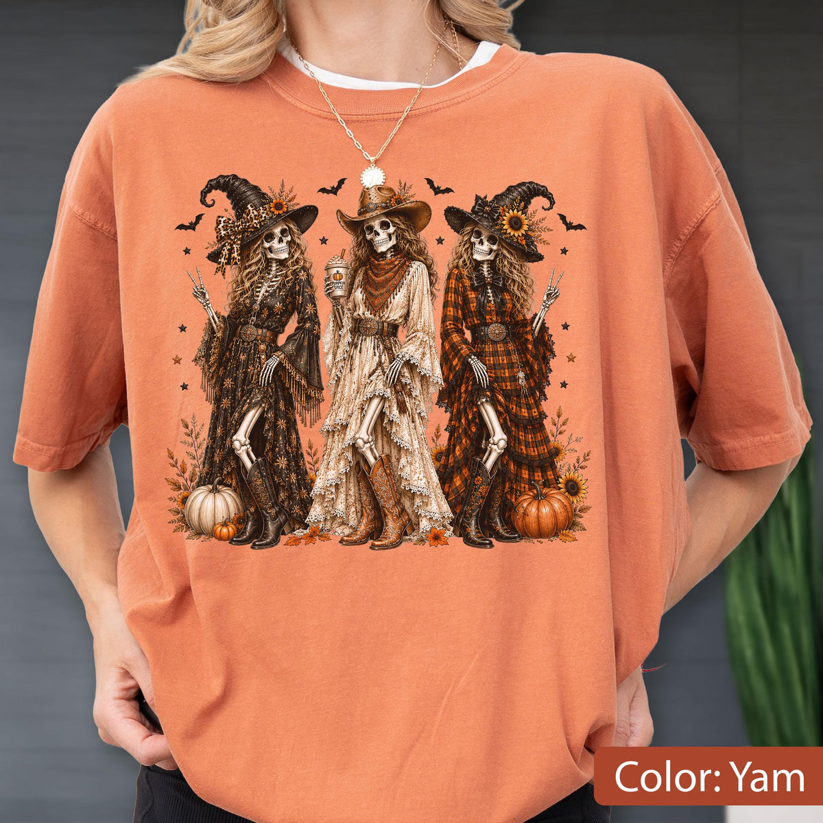 May include: A burnt orange t-shirt featuring a graphic print of three skeletons dressed in western-style witch attire, including hats, long dresses, and cowboy boots. The skeletons are surrounded by pumpkins, bats, and stars in a fall-themed design.