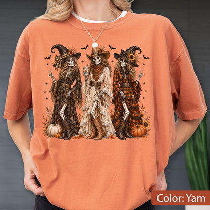 May include: A burnt orange t-shirt featuring a graphic print of three skeletons dressed in western-style witch attire, including hats, long dresses, and cowboy boots. The skeletons are surrounded by pumpkins, bats, and stars in a fall-themed design.