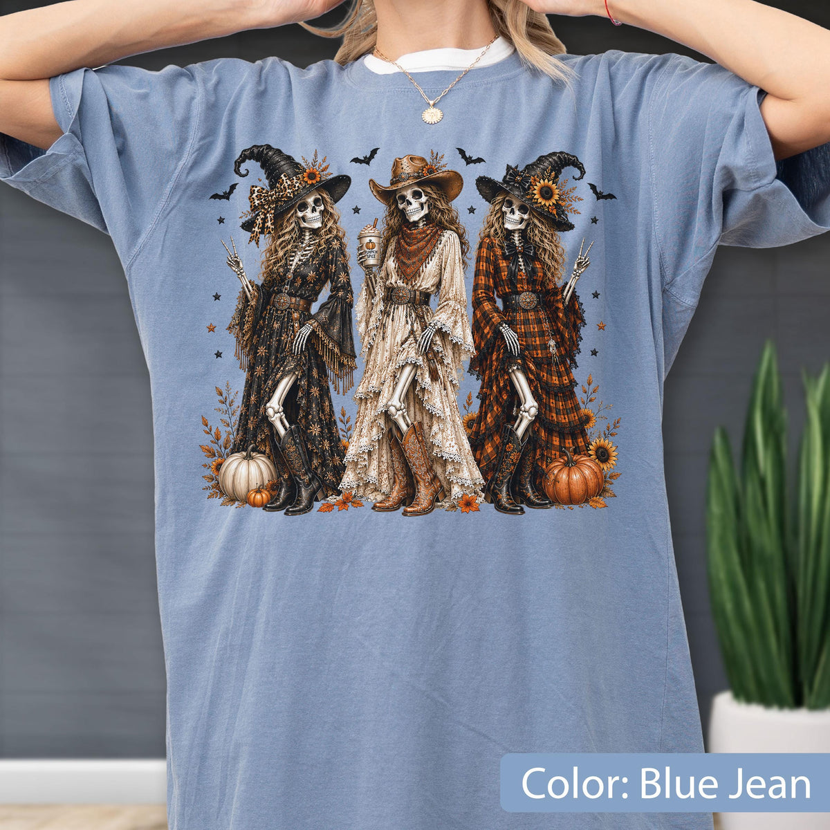 May include: A blue jean colored t-shirt featuring a graphic print of three skeletons dressed in western-style clothing, including hats, long dresses, and boots. The skeletons are surrounded by small pumpkins, sunflowers, and bats.