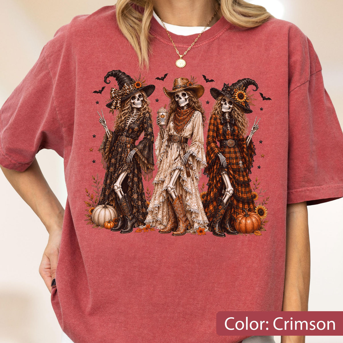 May include: A crimson short-sleeve t-shirt featuring a graphic of three skeletons dressed in western-style clothing, including hats and boots. The skeletons are surrounded by pumpkins, sunflowers, and bats. The text Color: Crimson is visible.
