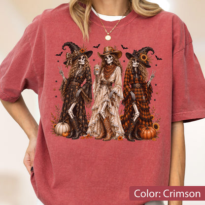 May include: A crimson short-sleeve t-shirt featuring a graphic of three skeletons dressed in western-style clothing, including hats and boots. The skeletons are surrounded by pumpkins, sunflowers, and bats. The text Color: Crimson is visible.
