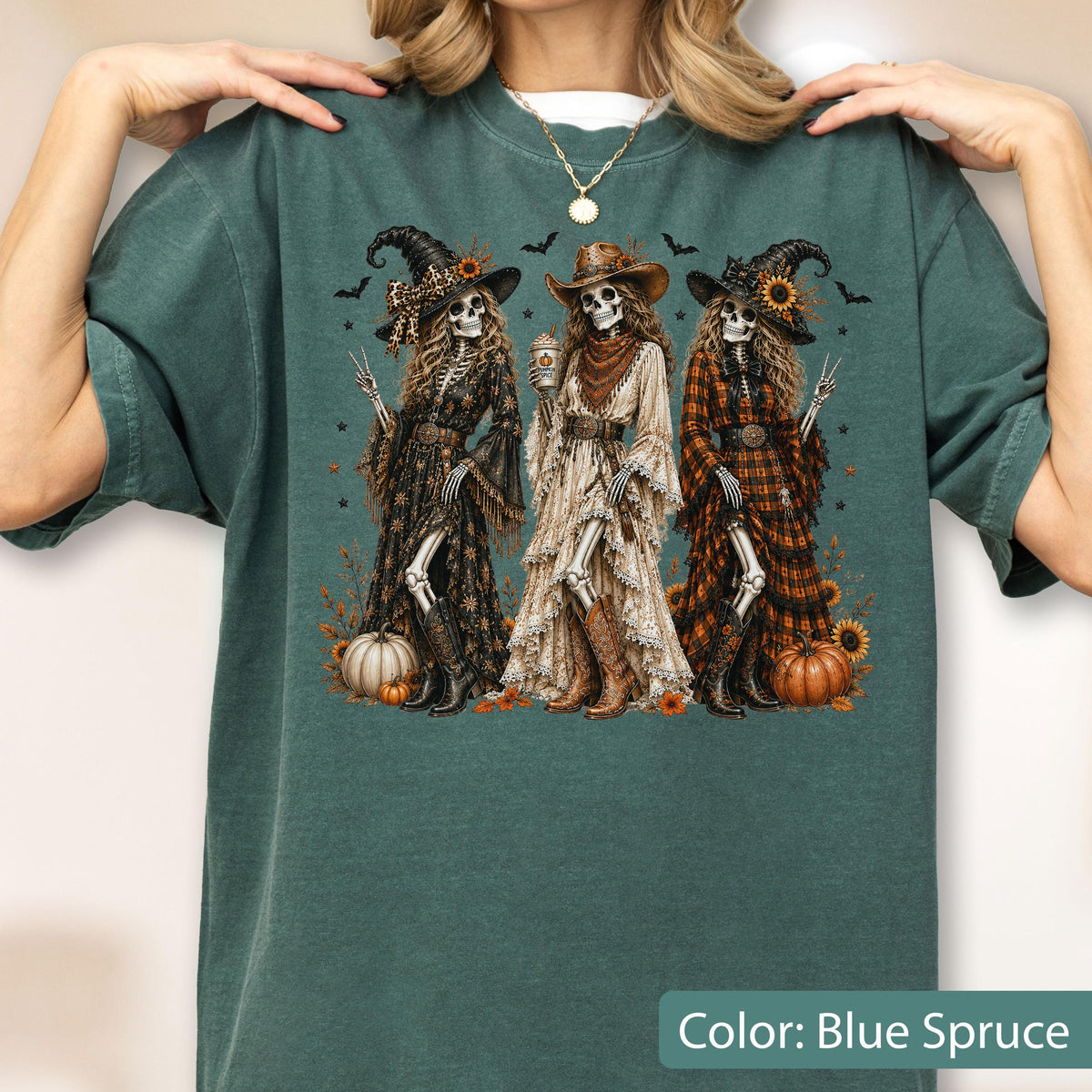 May include: A blue spruce t-shirt featuring a graphic of three skeletons dressed as witches in western-style clothing, including hats, dresses, and boots. The skeletons are surrounded by sunflowers, pumpkins, and small bats in a fall-themed design.