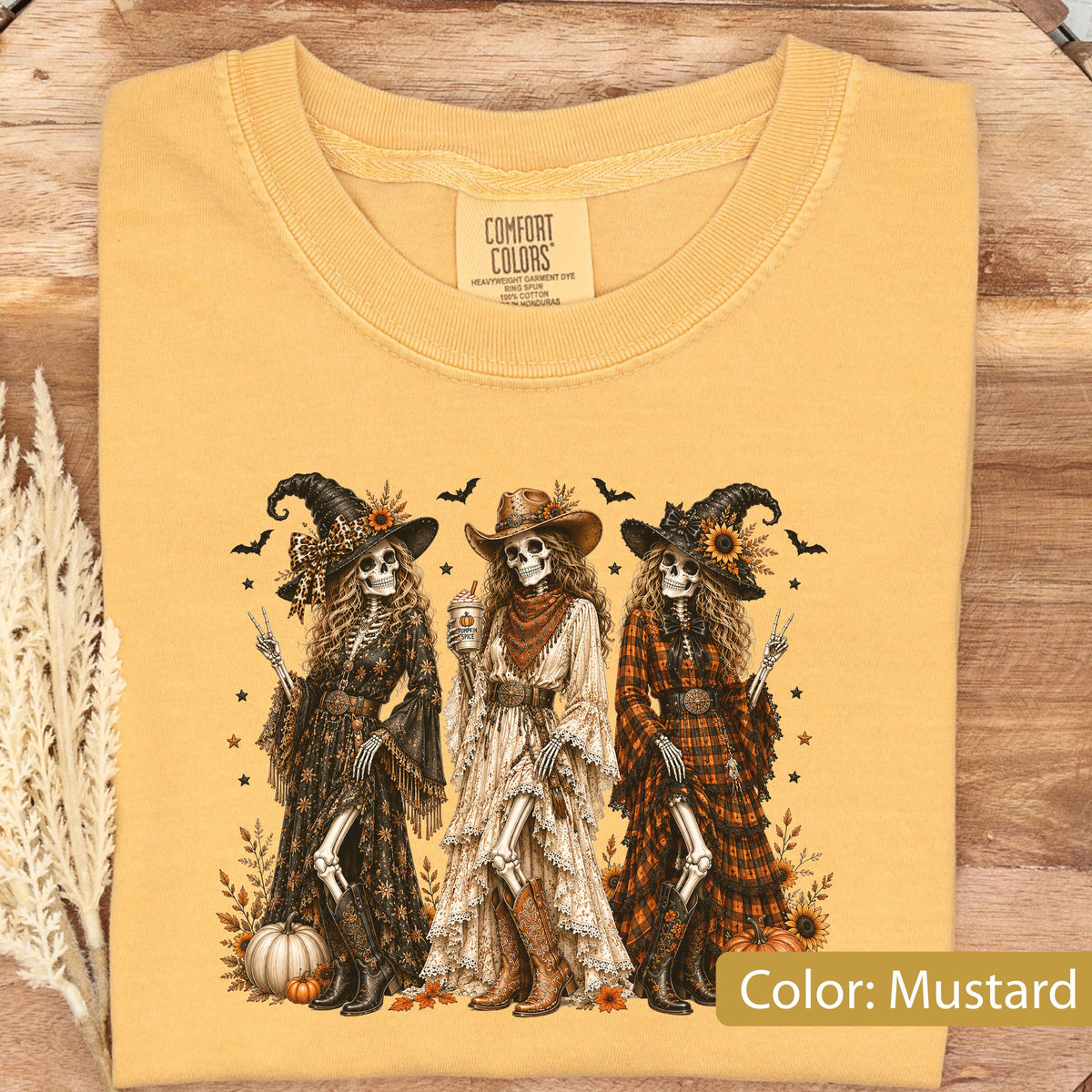 May include: A mustard yellow t-shirt featuring a graphic of three skeletons dressed in western-style witch attire. The skeletons wear hats, long dresses, and boots, surrounded by pumpkins, sunflowers, bats, and stars. The inner label reads Comfort Colors.