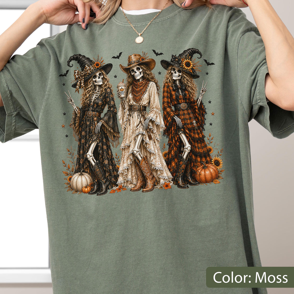 May include: A moss green t-shirt featuring a graphic print of three skeletons dressed as witches in western-style clothing, including hats, dresses, and boots. The design includes sunflowers, pumpkins, and bats, with the skeletons posing together.