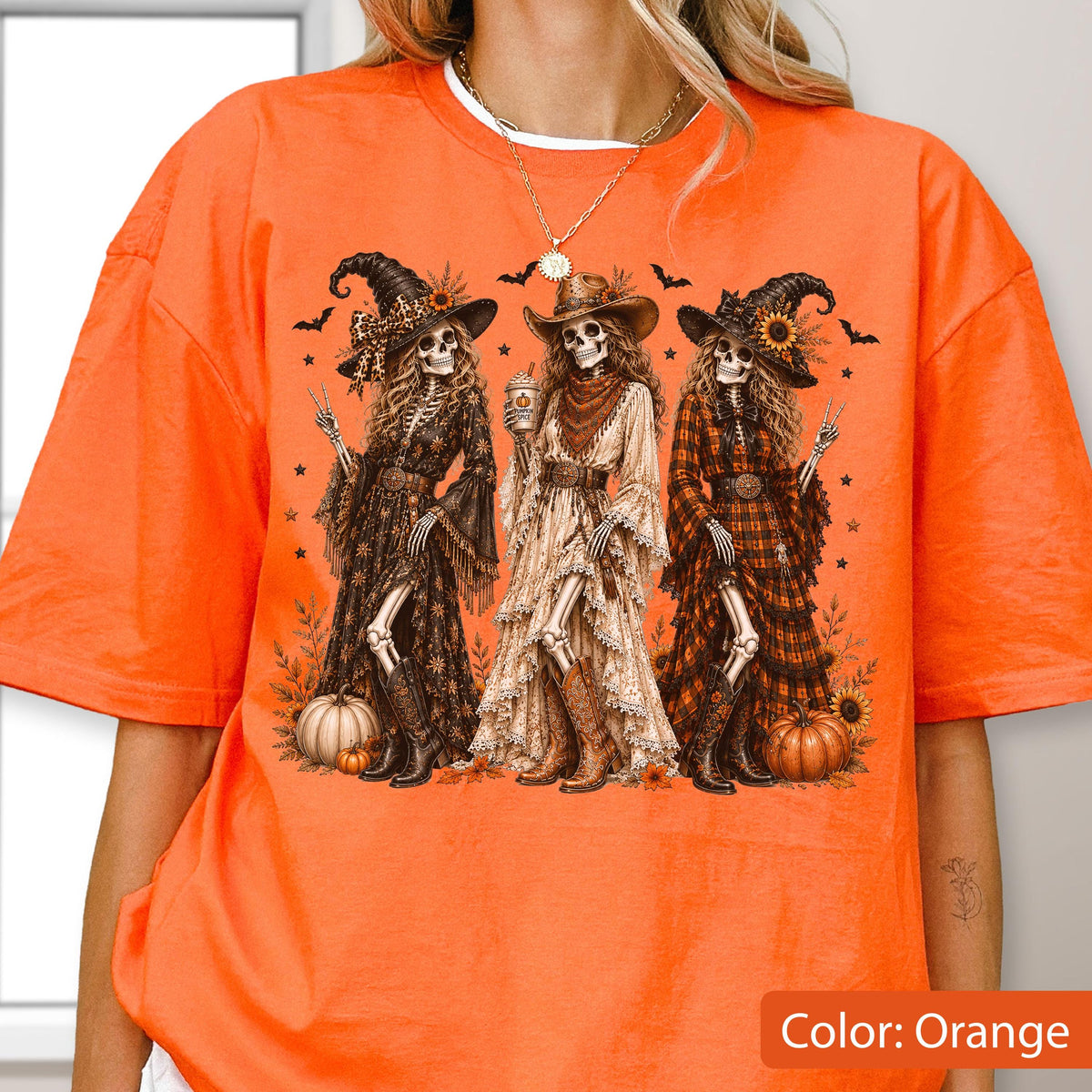 May include: An orange t-shirt featuring a graphic print of three skeletons dressed as witches and cowgirls. The skeletons wear hats, long dresses, and boots, surrounded by pumpkins, sunflowers, and bats. The design includes stars and foliage.
