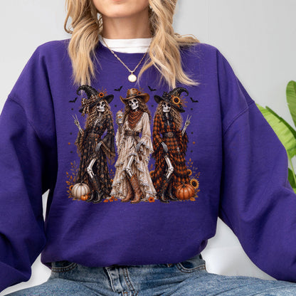 May include: A purple long-sleeve sweatshirt featuring a graphic print of three skeleton figures dressed as witches and cowgirls. The skeletons wear hats, dresses, and boots, surrounded by small pumpkins, sunflowers, and bats against a dark background.