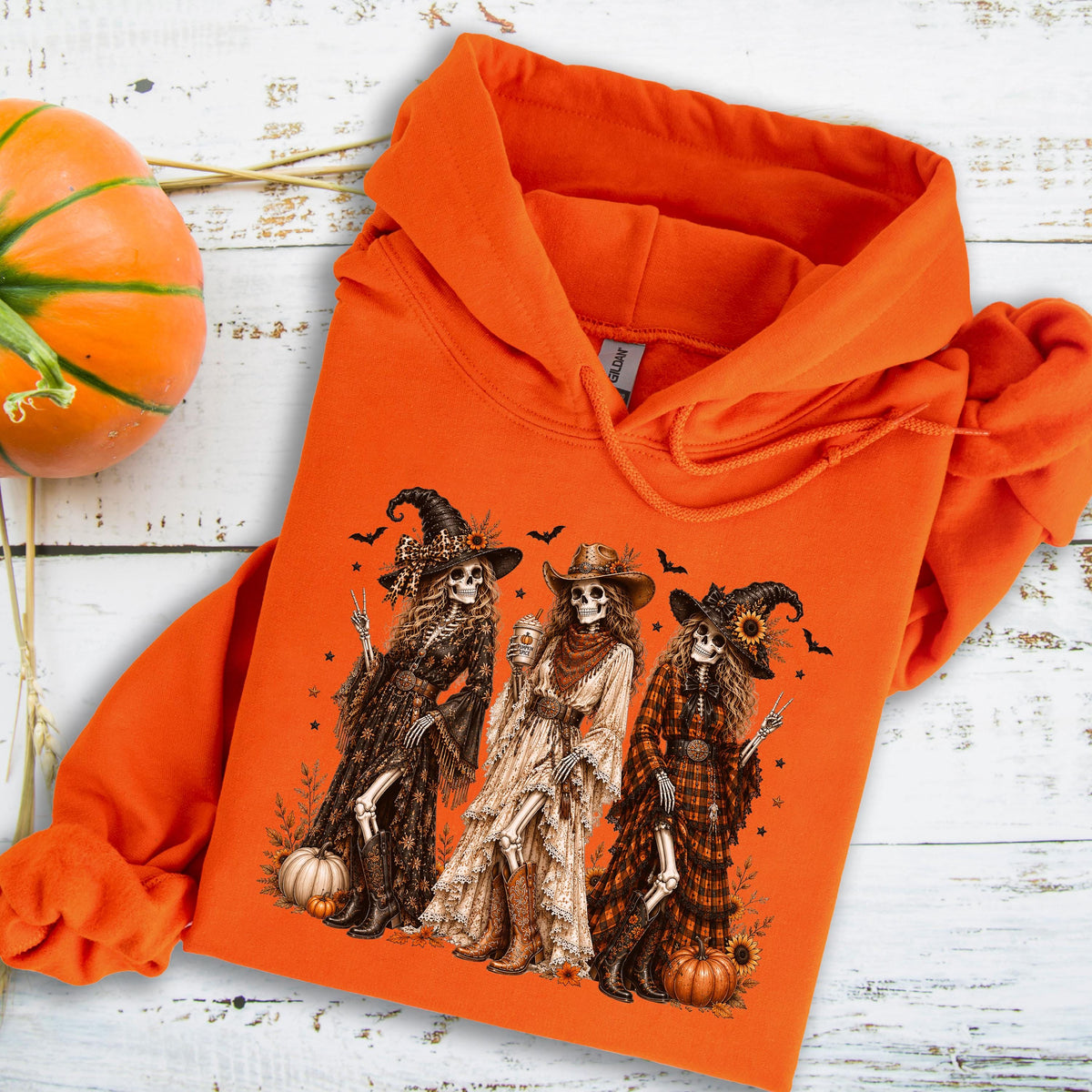 May include: An orange hooded sweatshirt featuring a graphic print of three skeletons dressed in western-style clothing, including hats and boots, surrounded by small bats and pumpkins. The hoodie has a drawstring hood and long sleeves.