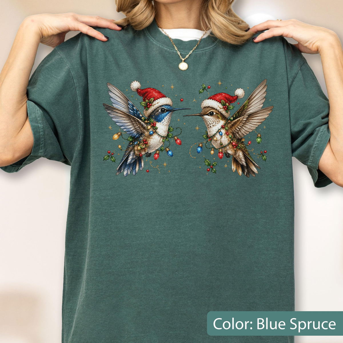 May include: A blue spruce colored t-shirt featuring a graphic of two hummingbirds wearing red Santa hats. The birds are surrounded by colorful Christmas light bulbs, holly leaves, and berries. The shirt has a crew neckline and short sleeves.