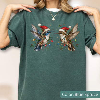 May include: A blue spruce colored t-shirt featuring a graphic of two hummingbirds wearing red Santa hats. The birds are surrounded by colorful Christmas light bulbs, holly leaves, and berries. The shirt has a crew neckline and short sleeves.