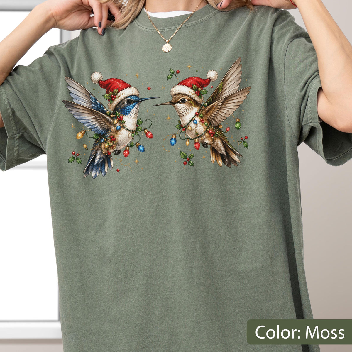 May include: A moss green short-sleeve t-shirt featuring a graphic print of two hummingbirds wearing red Santa hats. The birds are surrounded by colorful Christmas light bulbs and sprigs of holly. The shirt has a crew neckline and a relaxed fit.