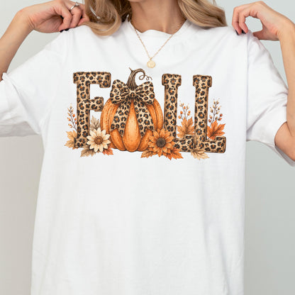 May include: A white t-shirt featuring a graphic print of the word FALL in leopard print lettering. An orange pumpkin with a leopard print bow sits in the center, surrounded by brown and yellow autumn leaves and sunflowers at the base.