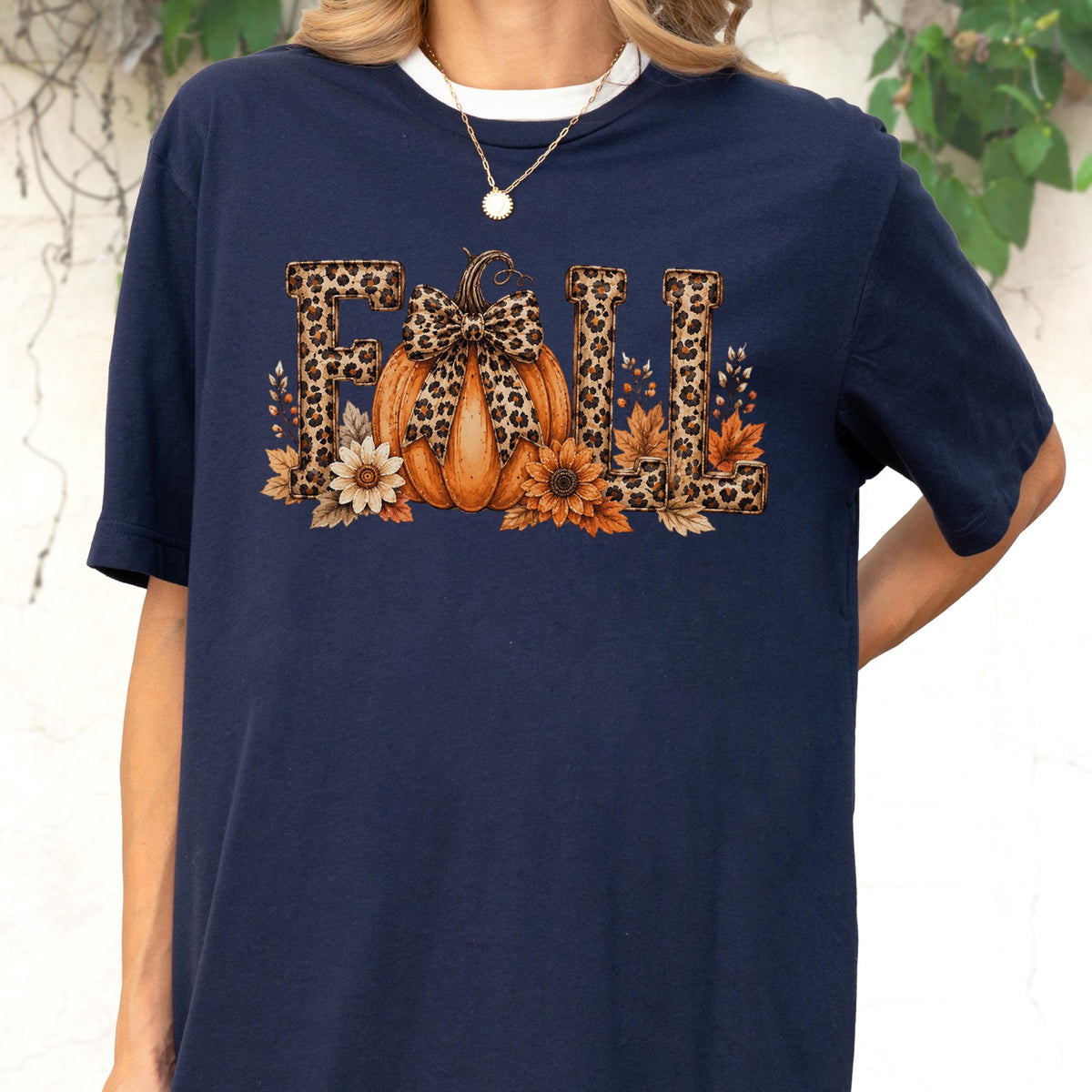 May include: A navy blue short-sleeve t-shirt featuring a graphic print of the word FALL. The letters have a leopard print pattern. An orange pumpkin with a leopard print bow sits in the center, surrounded by sunflowers and autumn leaves.