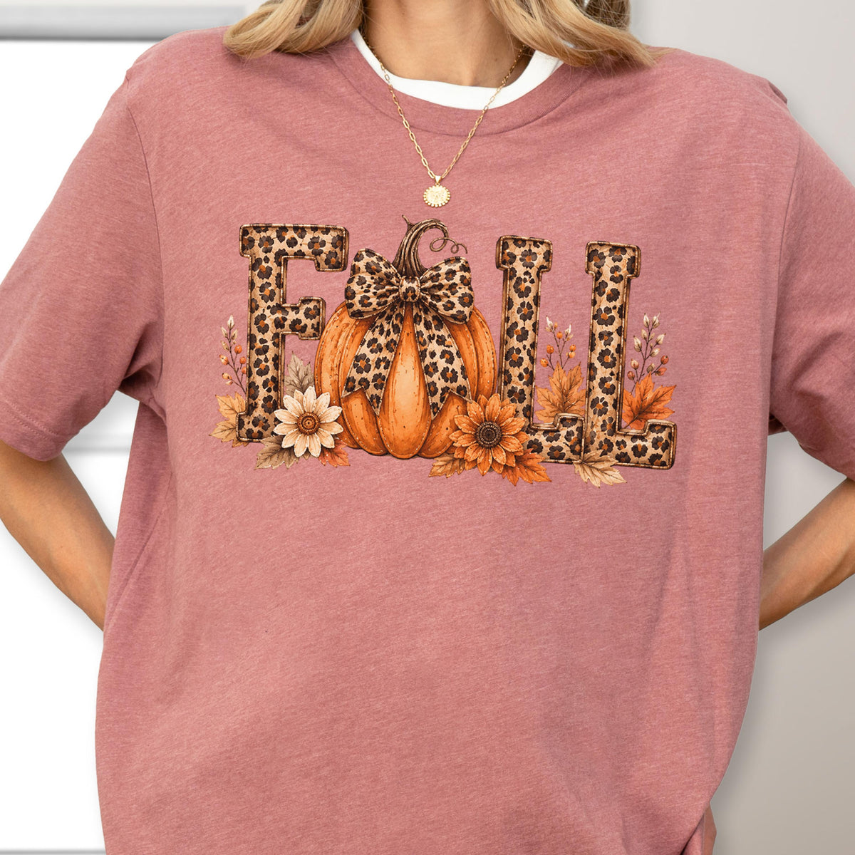 May include: A mauve short-sleeved t-shirt featuring a graphic print of the word FALL in a leopard print pattern. The letter A is replaced by an orange pumpkin with a leopard print bow, surrounded by sunflowers and autumn leaves.