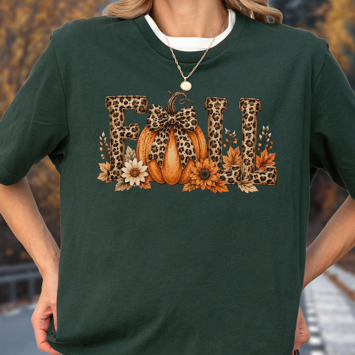 May include: A dark green short-sleeve t-shirt featuring a graphic print of the word FALL with leopard print lettering. An orange pumpkin with a leopard print bow sits in the center, surrounded by sunflowers and autumn leaves in shades of brown.