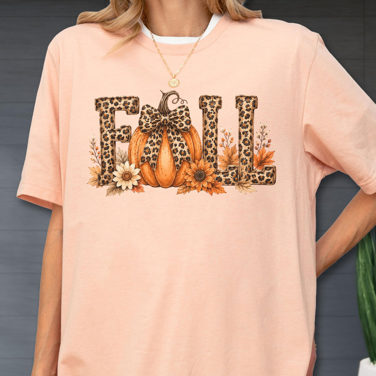 May include: A peach-colored short-sleeve t-shirt featuring a graphic print of the word FALL. The letters are filled with a leopard print pattern. An orange pumpkin with a leopard print bow sits in the center, surrounded by sunflowers and leaves.