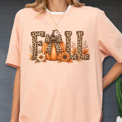 May include: A peach-colored short-sleeve t-shirt featuring a graphic print of the word FALL. The letters are filled with a leopard print pattern. An orange pumpkin with a leopard print bow sits in the center, surrounded by sunflowers and leaves.
