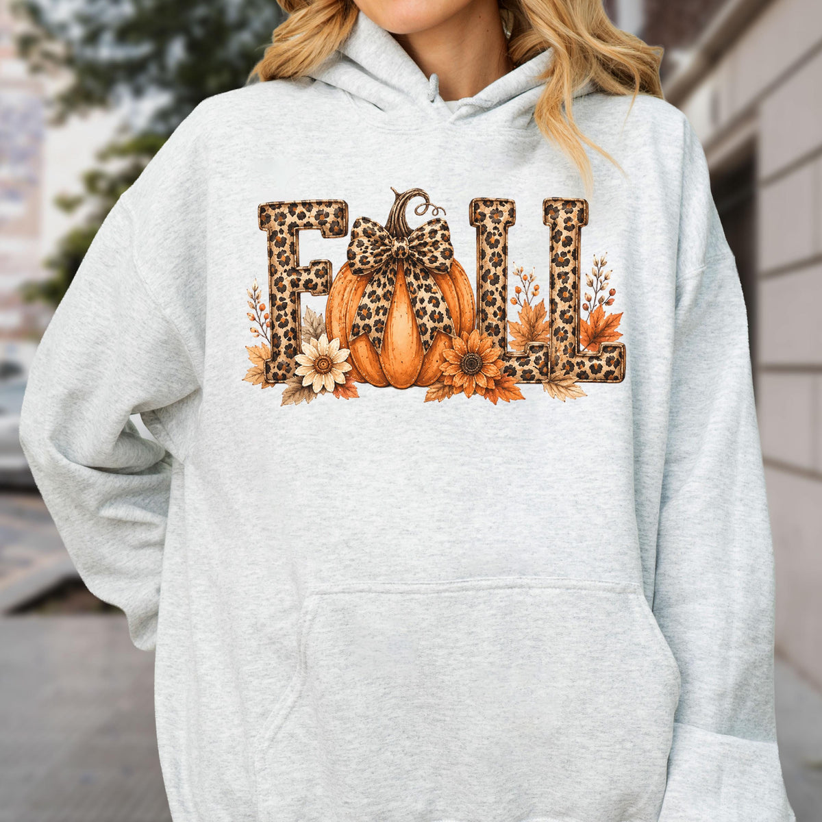 May include: A light gray hooded sweatshirt featuring a graphic print of the word FALL in leopard print lettering. An orange pumpkin with a leopard print bow sits in the center, surrounded by sunflowers and autumn leaves in shades of brown.
