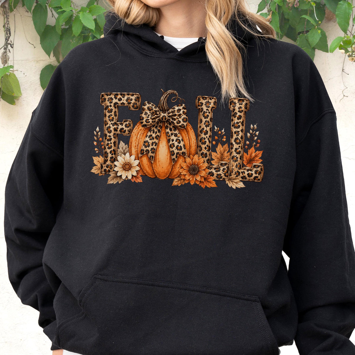 May include: A black hooded sweatshirt featuring a graphic print on the chest. The design displays the word FALL in leopard print lettering, with an orange pumpkin wearing a leopard print bow in the center, surrounded by sunflowers and leaves.