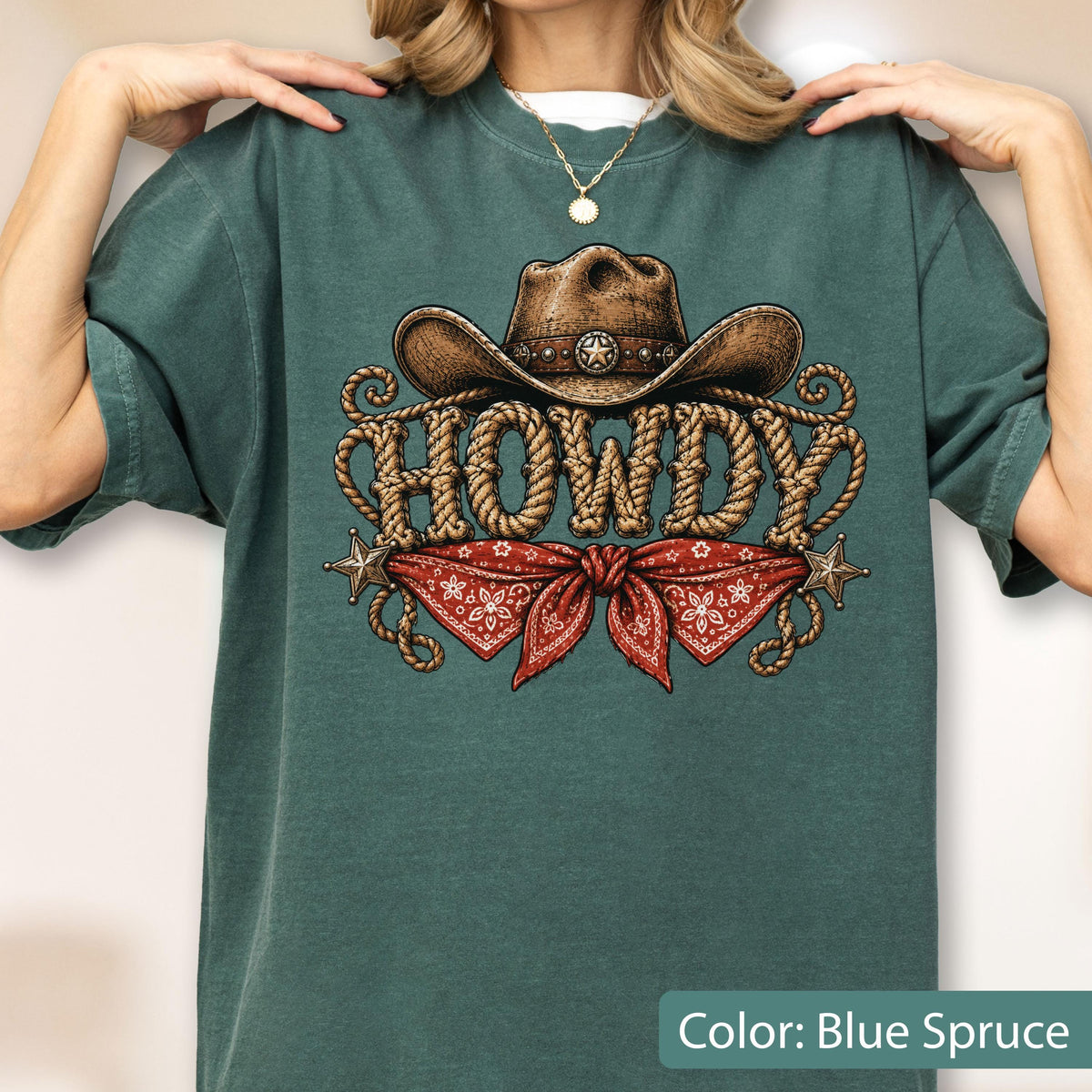 May include: A blue spruce t-shirt featuring a graphic print of a brown cowboy hat above the word HOWDY written in a rope-style font. Below the text is a red bandana with a white floral pattern and two small metallic stars on the sides.
