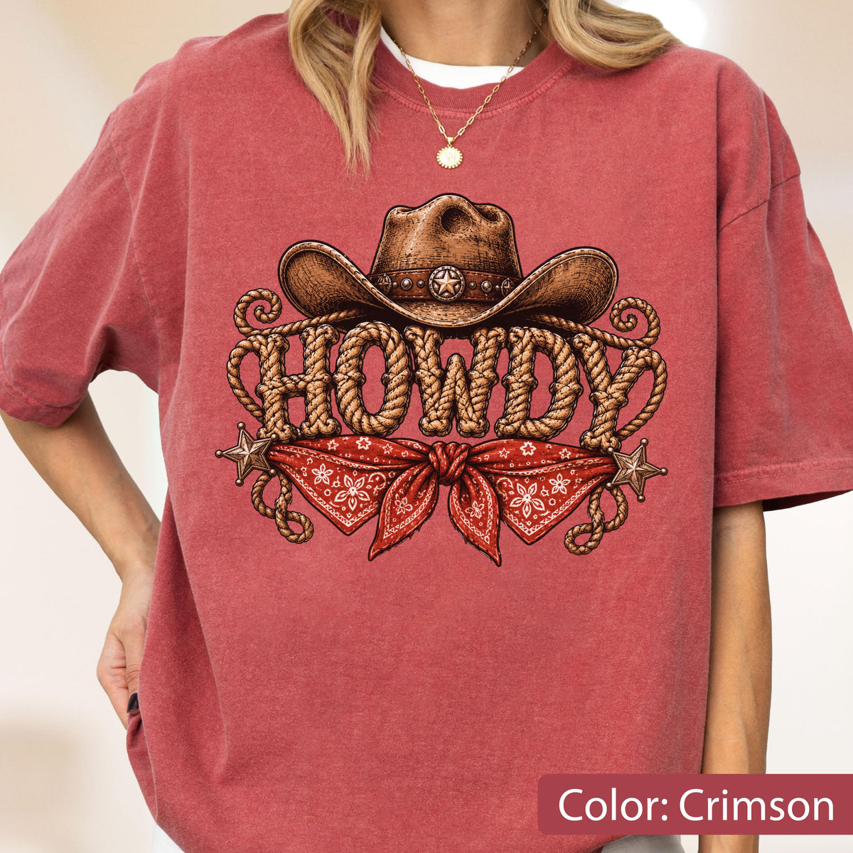 May include: A crimson short-sleeve t-shirt featuring a graphic print of a brown cowboy hat, a red bandana, and the word HOWDY written in a rope-style font. The design includes two small star accents on the left and right sides of the graphic.