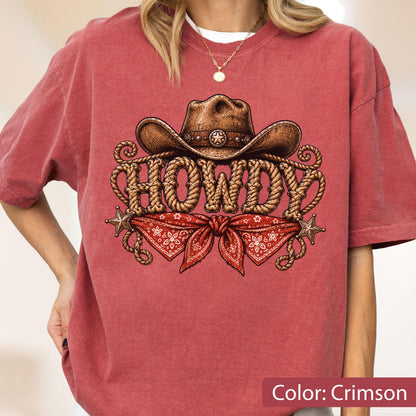 May include: A crimson short-sleeve t-shirt featuring a graphic print of a brown cowboy hat, a red bandana, and the word HOWDY written in a rope-style font. The design includes two small star accents on the left and right sides of the graphic.
