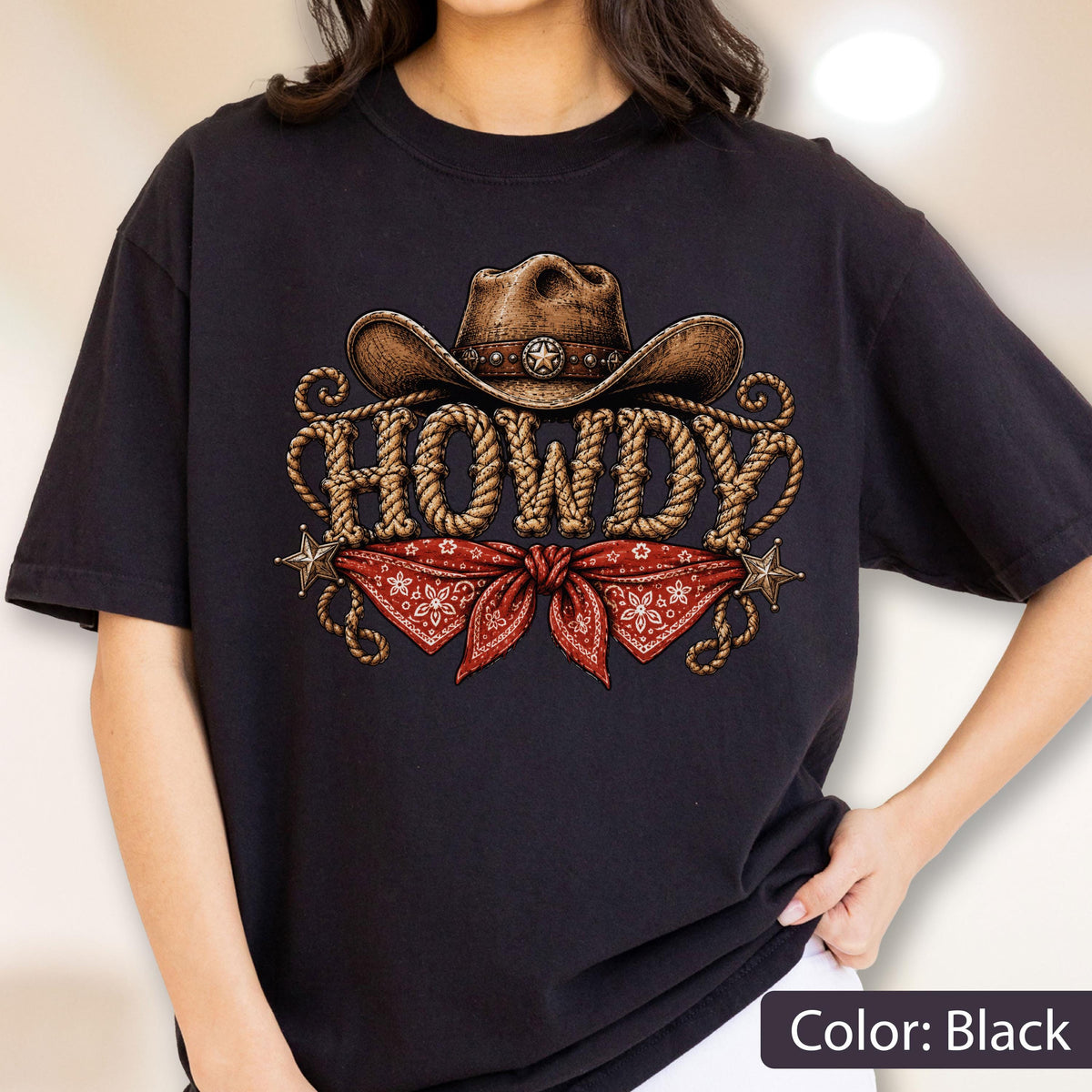 May include: A black t-shirt featuring a graphic print of a brown cowboy hat above the word HOWDY written in a rope-style font. Below the text is a red bandana with a white floral pattern, flanked by two small metallic-colored star icons.