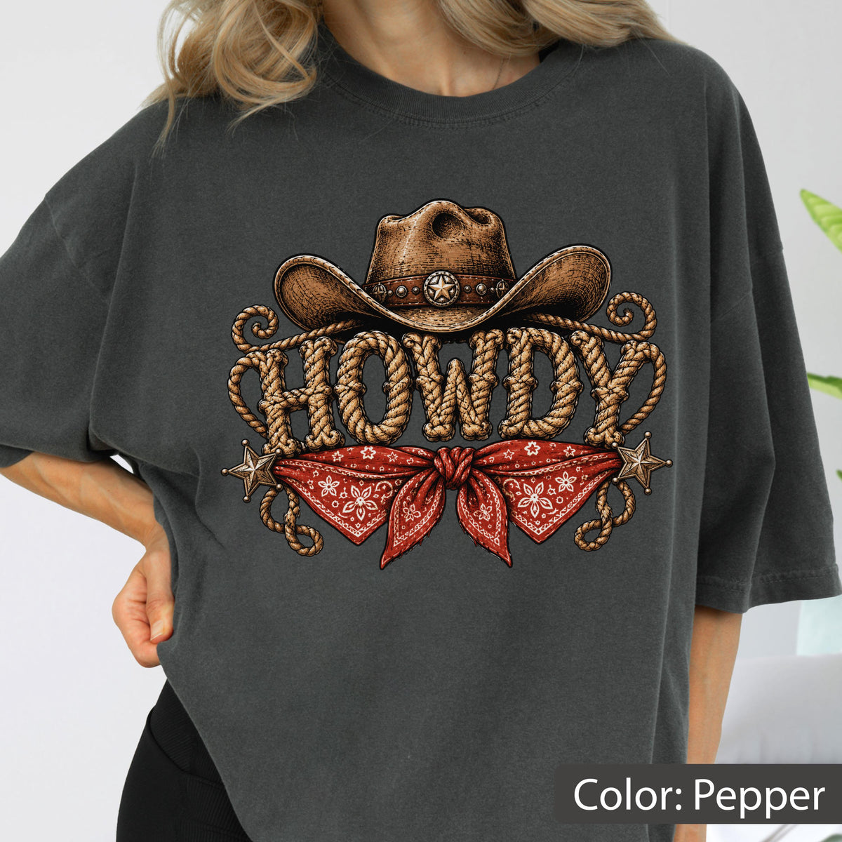 May include: A charcoal gray t-shirt featuring a graphic print of a brown cowboy hat above the word HOWDY written in a rope-style font. Below the text is a red bandana with a white floral pattern, flanked by two small brown stars.