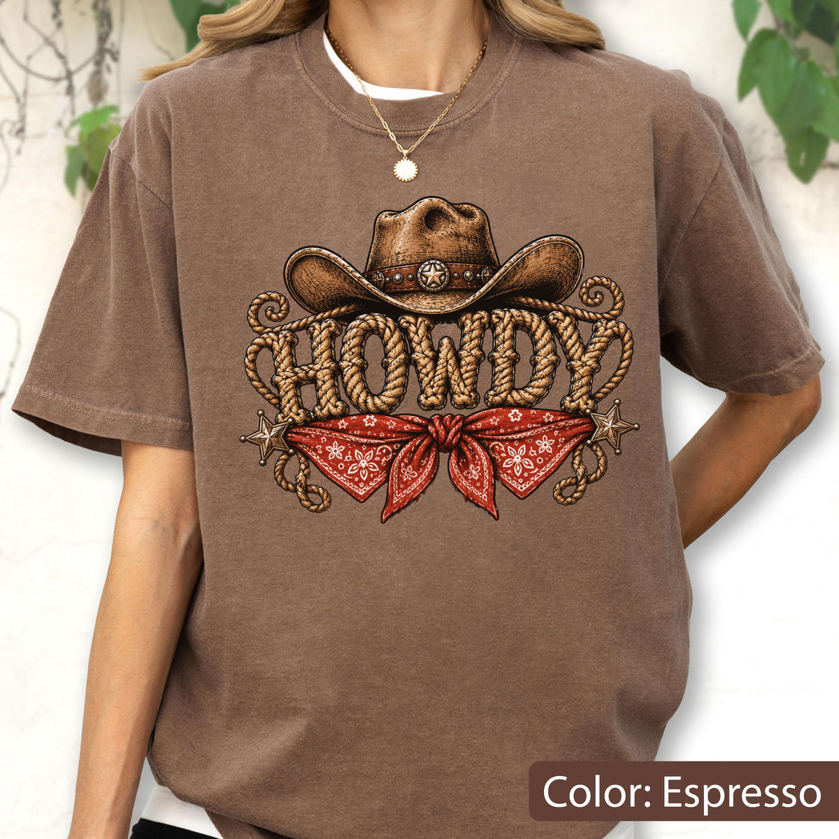 May include: An espresso-colored t-shirt featuring a graphic print of a brown cowboy hat above the word HOWDY written in a rope-style font, with a red patterned bandana tied below and two small star accents on the sides of the design.