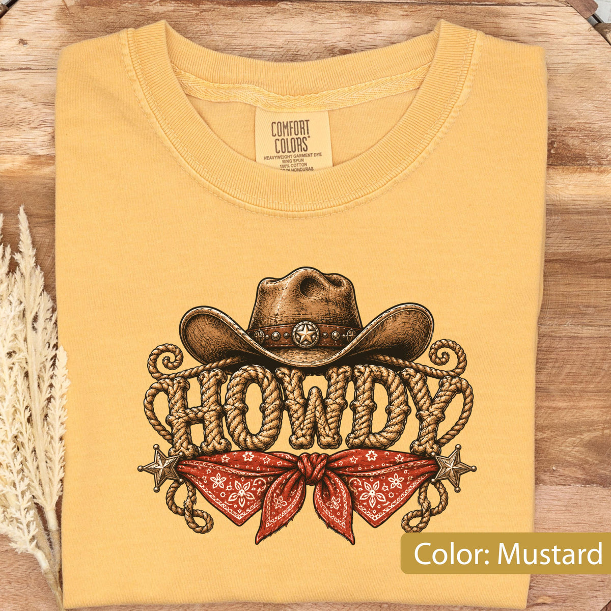 May include: A mustard yellow t-shirt featuring a graphic print of a brown cowboy hat resting above the word HOWDY, which is stylized to look like coiled rope. Below the text is a red bandana with a white pattern and two small silver stars.