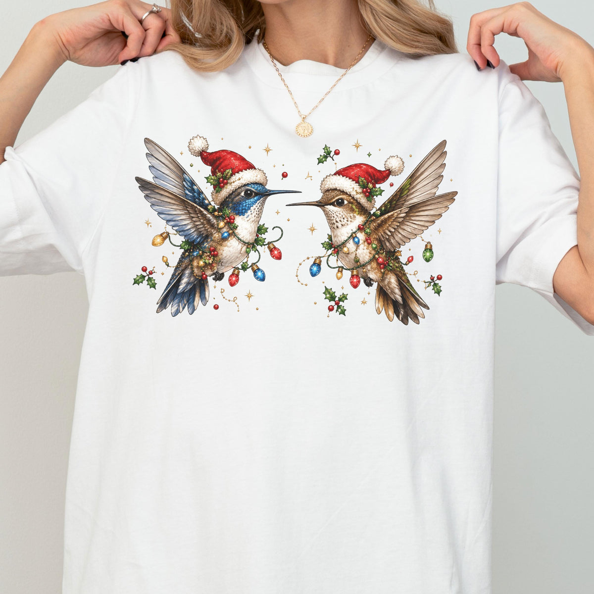 May include: A white short-sleeve t-shirt featuring a graphic print of two hummingbirds wearing red Santa hats. The birds are surrounded by colorful Christmas light bulbs, green holly leaves, and red berries, creating a festive holiday scene.