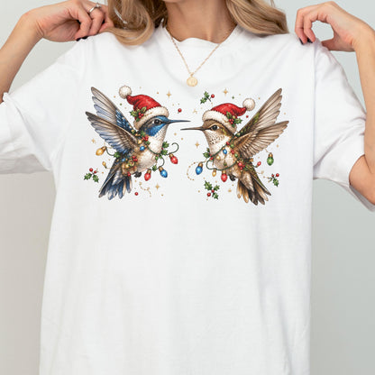 May include: A white short-sleeve t-shirt featuring a graphic print of two hummingbirds wearing red Santa hats. The birds are surrounded by colorful Christmas light bulbs, green holly leaves, and red berries, creating a festive holiday scene.