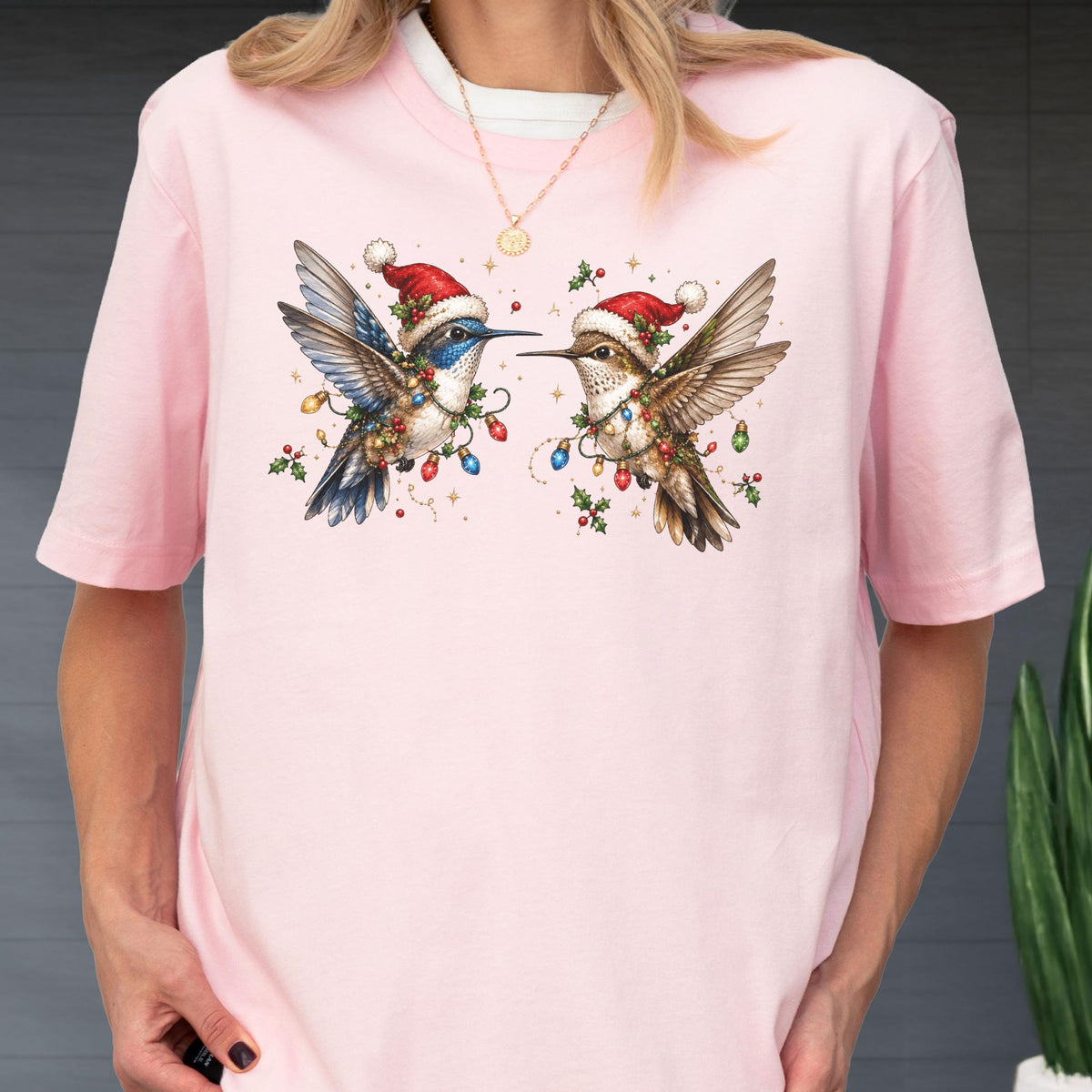 May include: A light pink short-sleeve t-shirt featuring a graphic print of two hummingbirds wearing red Santa hats. The birds are decorated with colorful Christmas lights and holly berries, with one bird showing blue plumage and the other brown.
