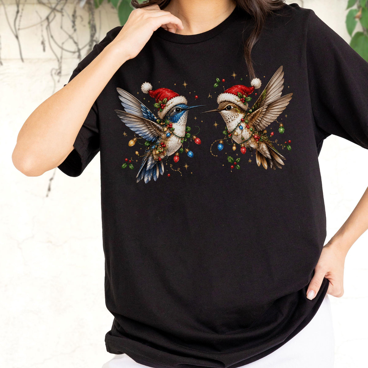May include: A black t-shirt featuring a graphic of two hummingbirds wearing red and white Santa hats. The birds are surrounded by colorful Christmas lights and holly berries, with one bird showing blue plumage and the other showing brown tones.