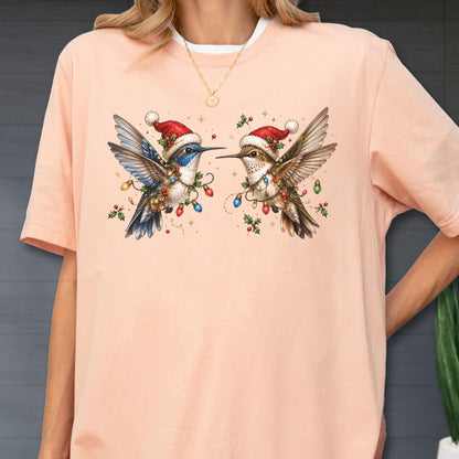 May include: A peach-colored short-sleeve t-shirt featuring a graphic of two hummingbirds facing each other. Both birds wear red and white Santa hats and are adorned with colorful Christmas light strings and green holly leaves with red berries.